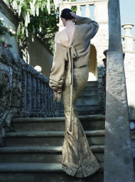Mariacarla Boscono climbs outdoor stone stairs wearing a beige tie-back sweater and a metallic gold floor-length skirt.
