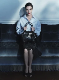 Mariacarla Boscono in a light blue shirt holding a black crocodile leather tote while seated on a tufted velvet sofa.