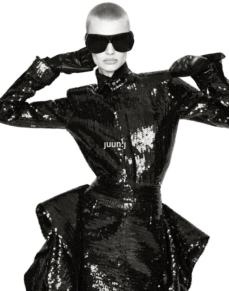 Model Mila van Eeten wearing a structured black sequin dress, dark oversized sunglasses, and leather gloves against a white studio backdrop.