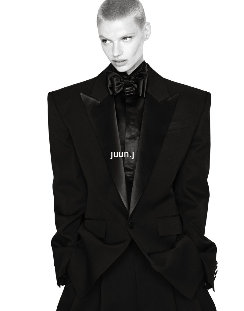 Model with a buzzcut wearing an oversized black tailored tuxedo jacket and bow tie against a white background for Juun.J F/W26