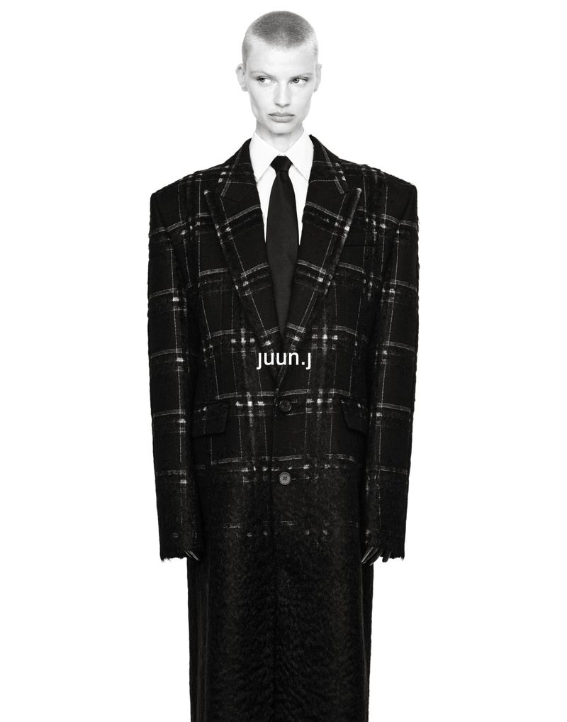 Model Mila van Eeten wearing an oversized black plaid tailored coat with a white shirt and black tie against a white backdrop.