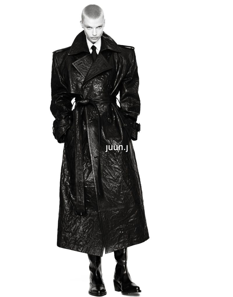 Model Mila van Eeten wearing a long belted black textured trench coat and leather boots against a solid white background.
