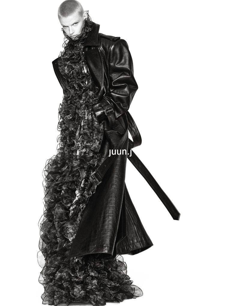 Model Mila van Eeten poses in an oversized black leather trench coat over a voluminous ruffled black dress on a white background.