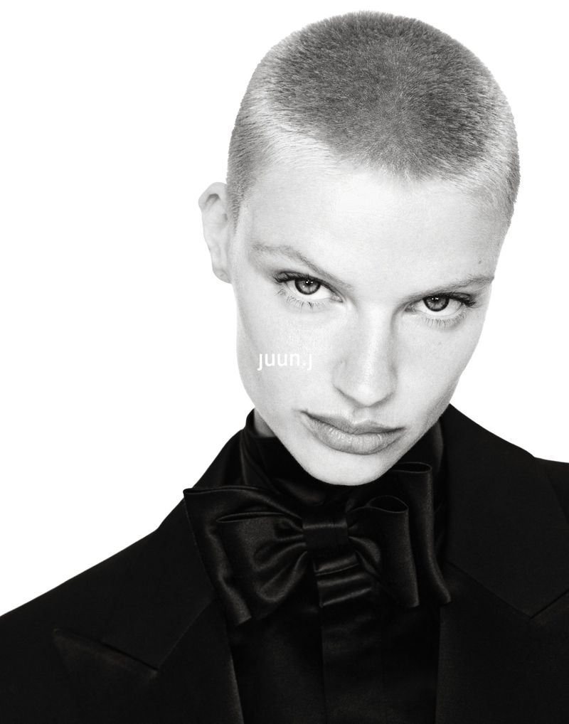 Mila van Eeten with a short buzz cut wearing a black tuxedo jacket and oversized satin bow tie against a white backdrop.