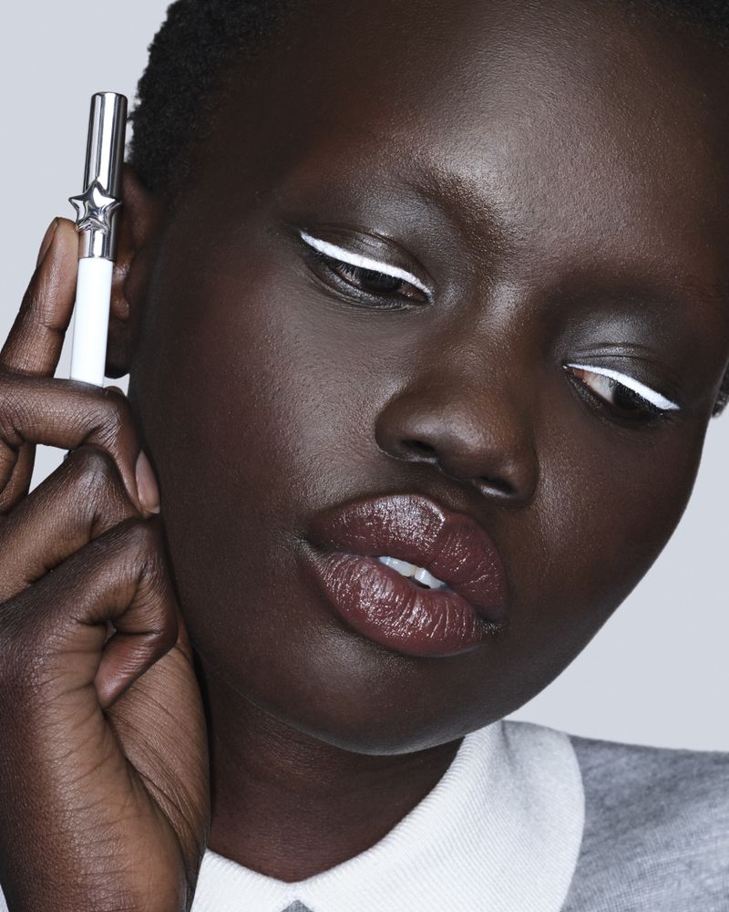 Close-up beauty portrait of model Aker Ajak wearing graphic white eyeliner and glossy lips while holding a cosmetic pencil.