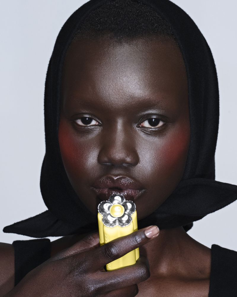 Model Aker Ajak wearing a black headscarf and holding a yellow Marc Jacobs Beauty floral lip product up to her lips.