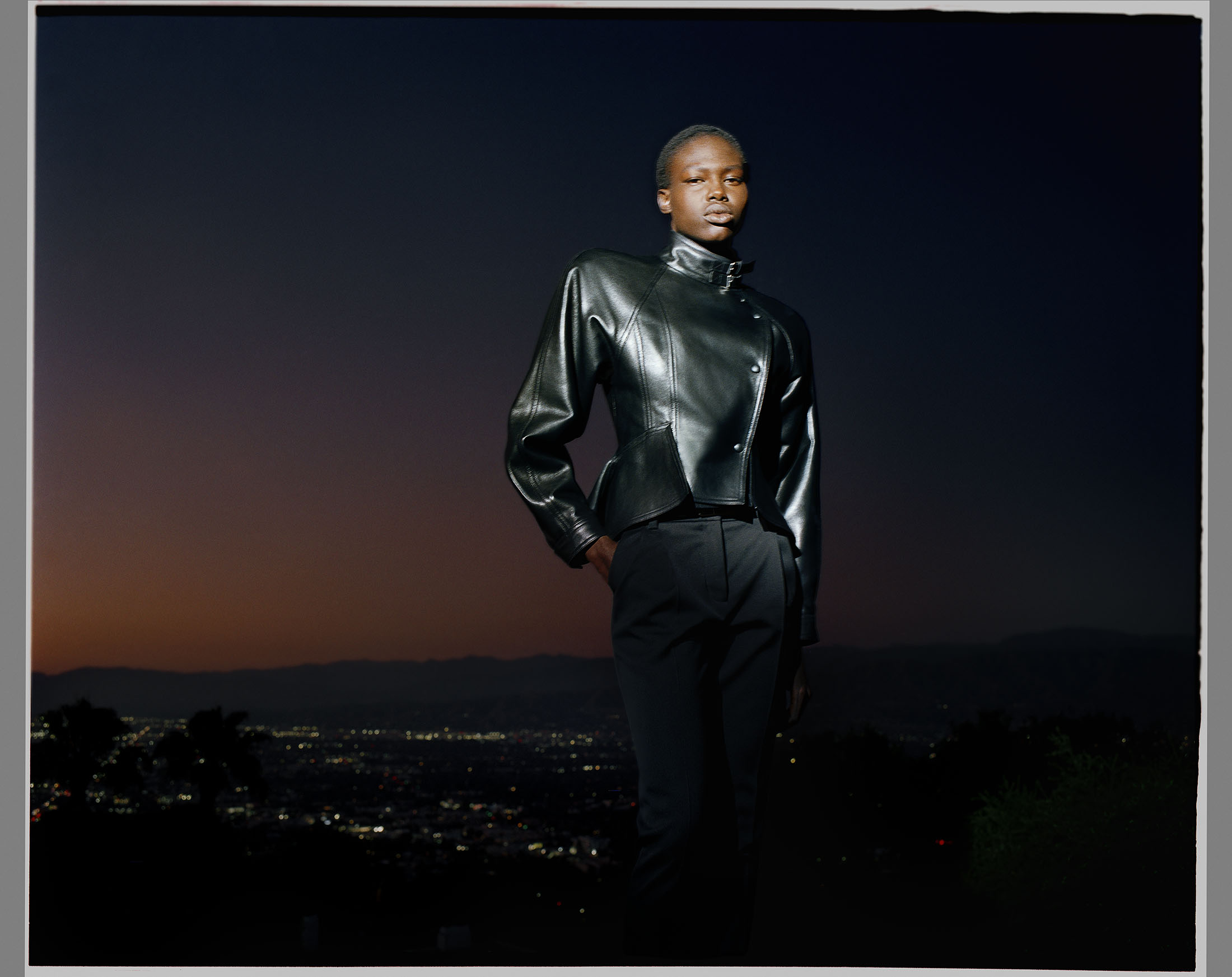 Model Abuk Yor poses outdoors at dusk wearing a structured black leather jacket and tailored trousers overlooking distant city lights.