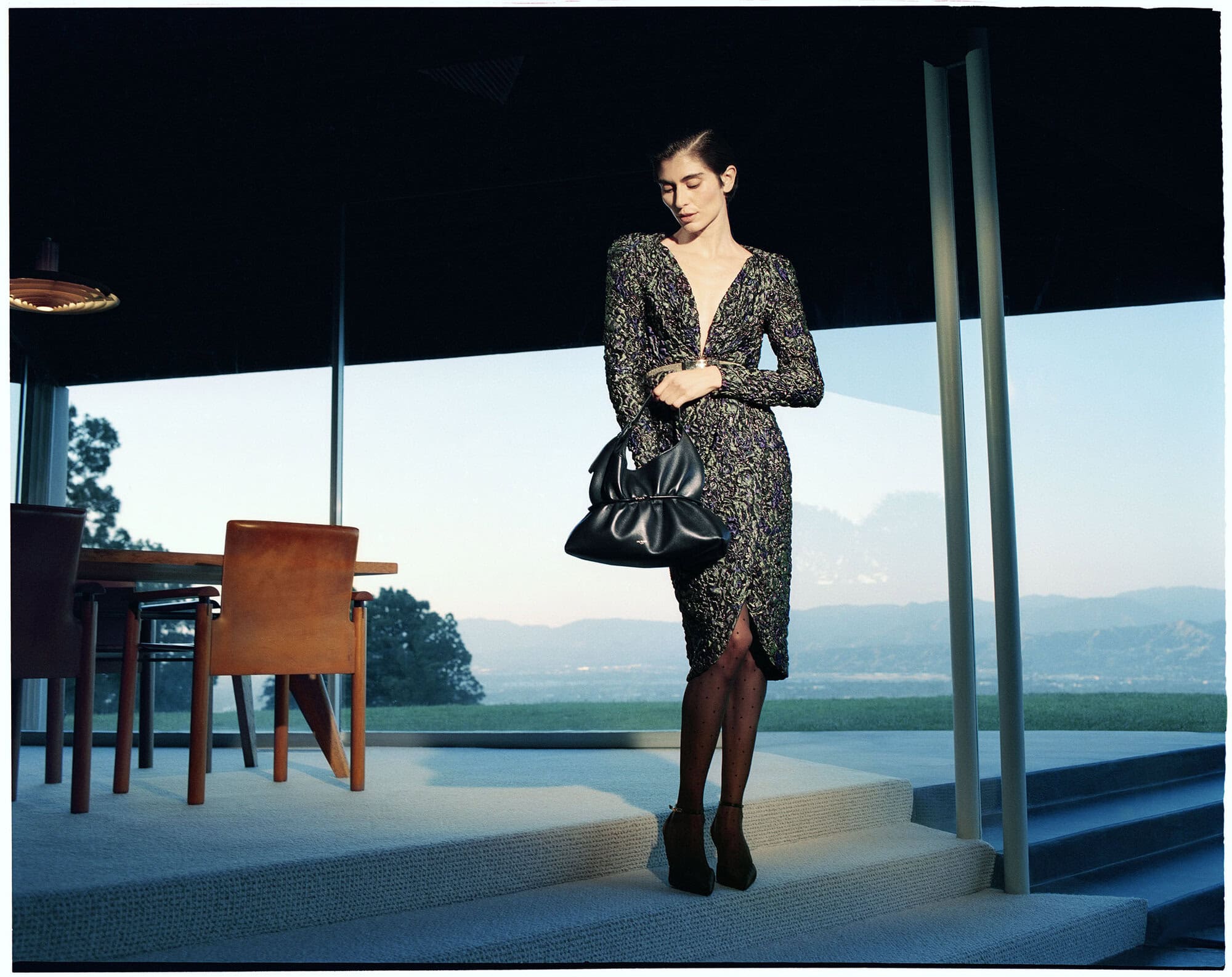 Model posing in a patterned deep-V dress holding a black leather handbag in a modernist glass pavilion at dusk.