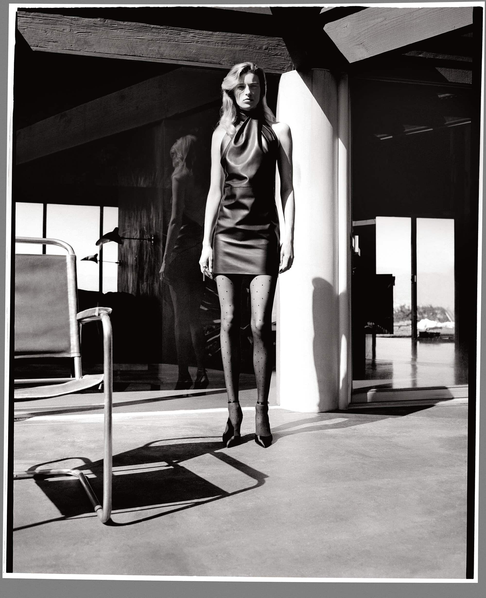 Black and white portrait of a model in a dark halter dress and polka dot tights standing in a modern interior.
