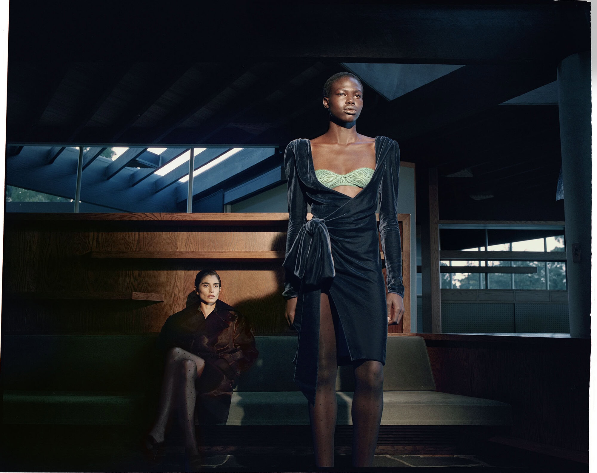 Model Abuk Yor standing in a draped black velvet dress with a seated model in the background of a modernist interior.