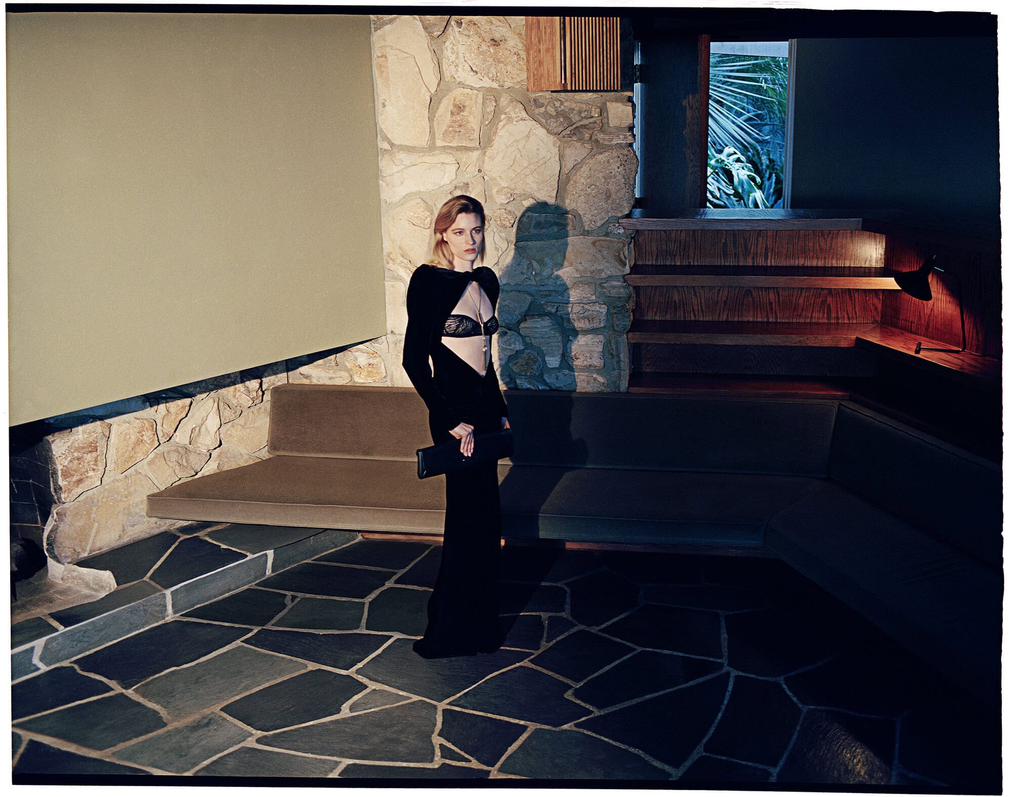 Model in a black cut-out evening gown holding a clutch standing on flagstone flooring in a modernist stone interior.