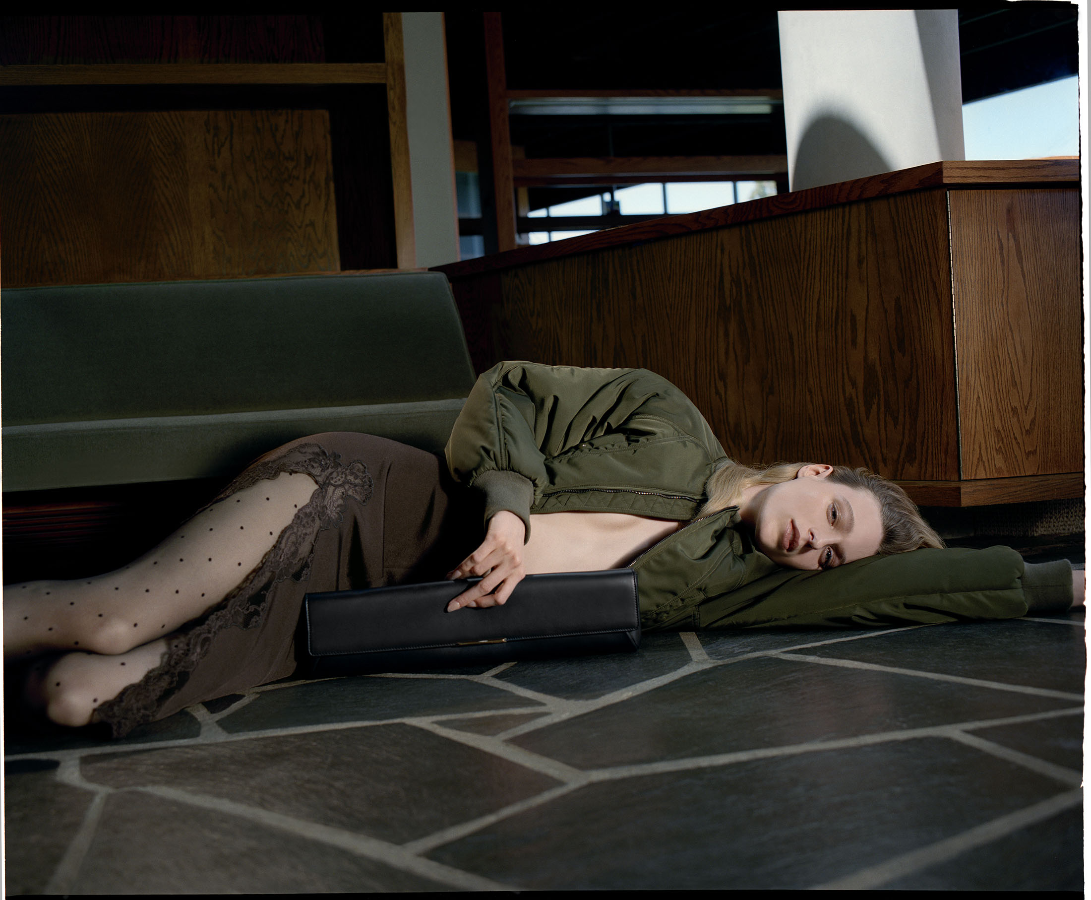 Model lying on a slate floor wearing an olive green bomber jacket, lace-trimmed skirt, and polka dot tights holding a black clutch.