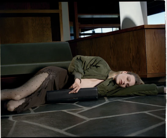 Model lying on a slate floor wearing an olive green bomber jacket, lace-trimmed skirt, and polka dot tights holding a black clutch.