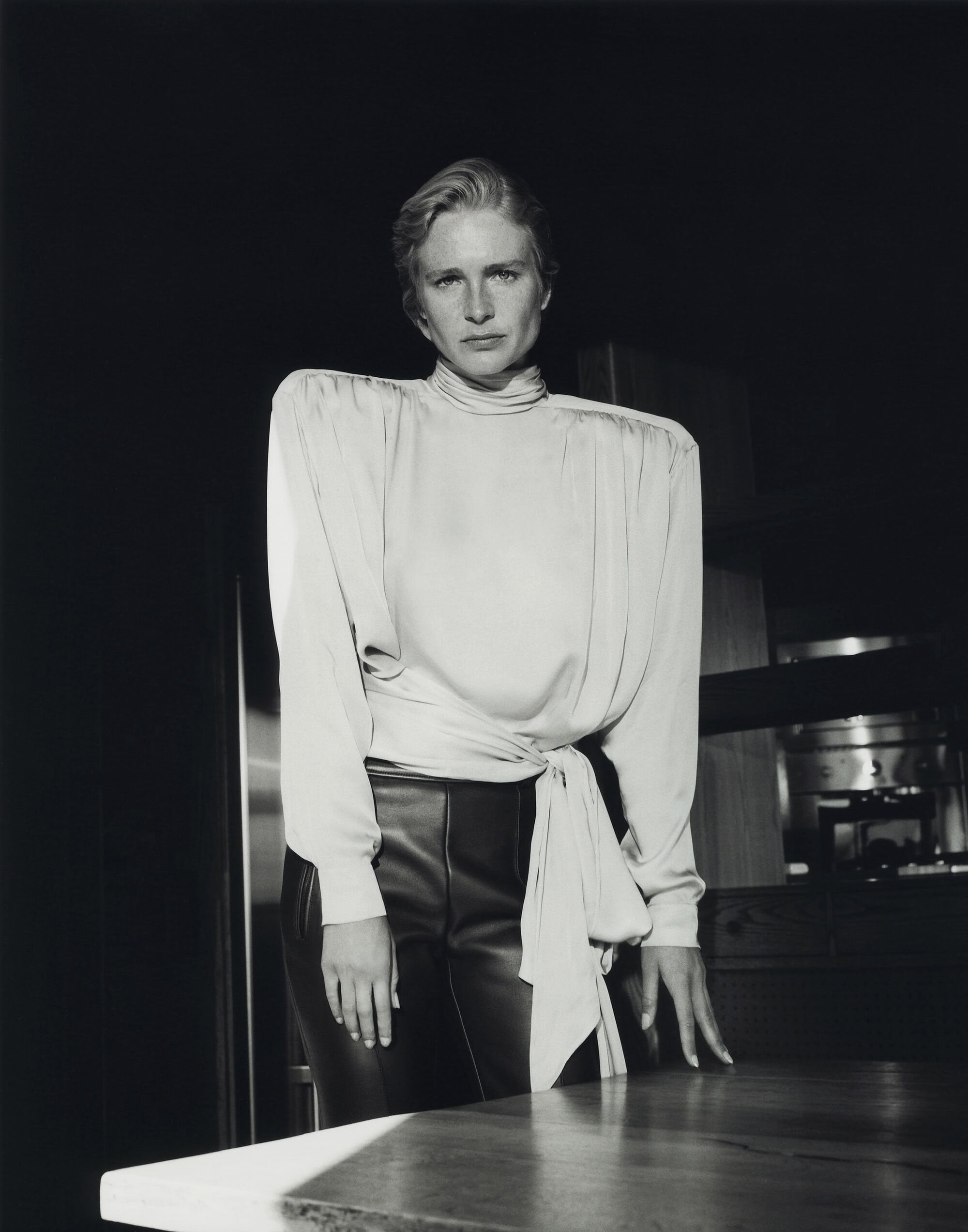 Model wearing a draped padded shoulder blouse and leather trousers posing in low light by a table.