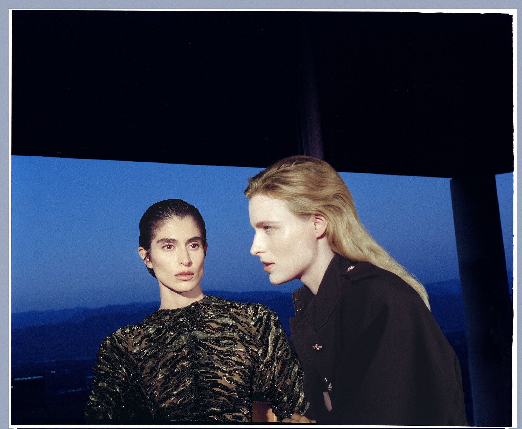Two female models posing indoors against a twilight landscape view for the Balmain Fall 2026 ad campaign.