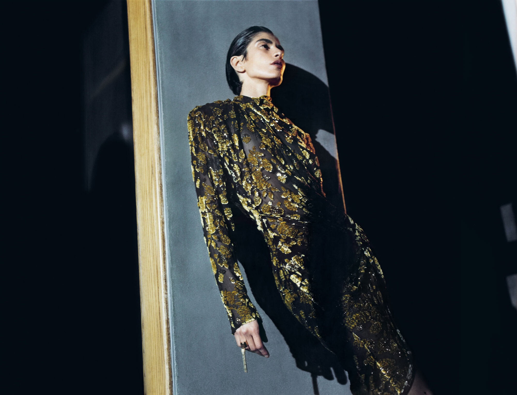 Fashion model in a metallic gold and black patterned long-sleeve dress leaning against a grey structural pillar in a dark room.