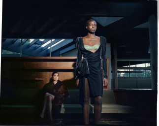 Model Abuk Yor standing in a draped black velvet dress with a seated model in the background of a modernist interior.