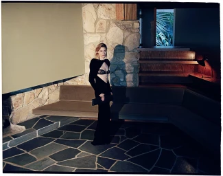 Model in a black cut-out evening gown holding a clutch standing on flagstone flooring in a modernist stone interior.