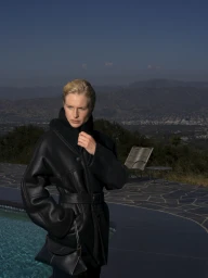 Model in black belted coat holding a black handbag by a pool overlooking distant mountains for Balmain