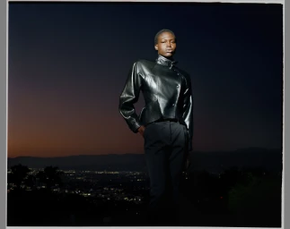 Model Abuk Yor poses outdoors at dusk wearing a structured black leather jacket and tailored trousers overlooking distant city lights.