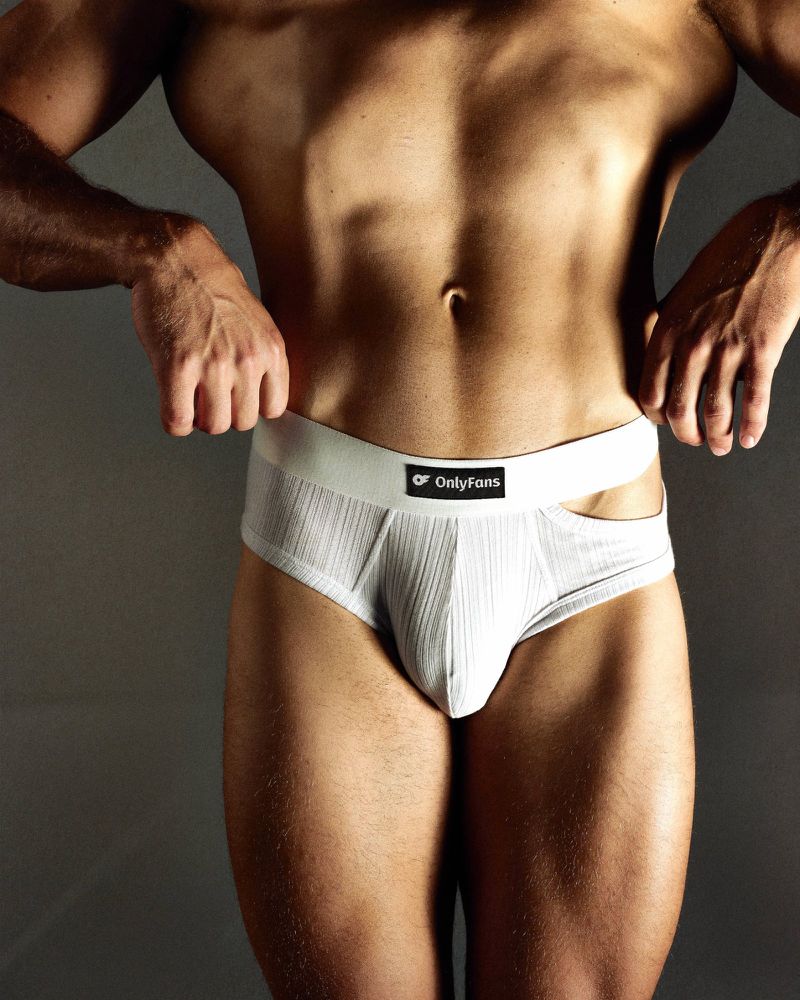 Cropped view of a muscular man wearing white ribbed briefs with an OnlyFans branded waistband against a dark studio backdrop.