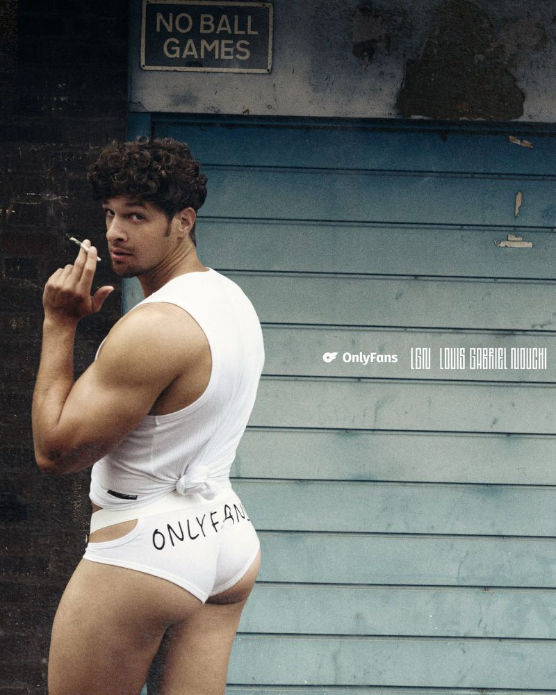 Man with curly hair looking over shoulder while smoking a cigarette, wearing a white tank top and OnlyFans underwear.