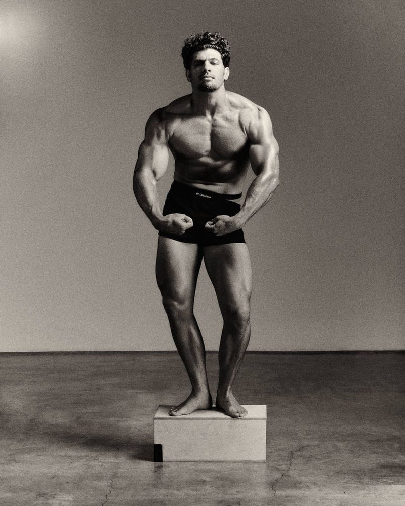 Muscular man posing and flexing on a wooden plinth wearing black briefs in a black and white studio setting.