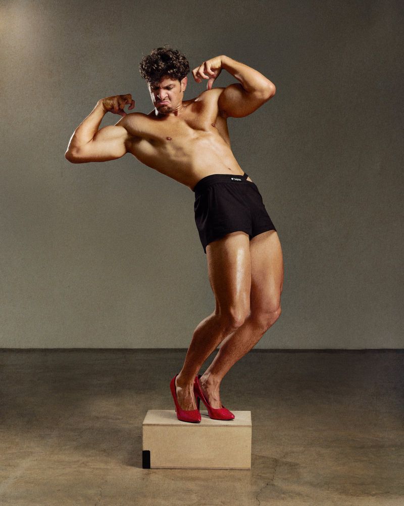 Shirtless man wearing black shorts and red high heels flexing muscles on a wooden block against a grey studio wall