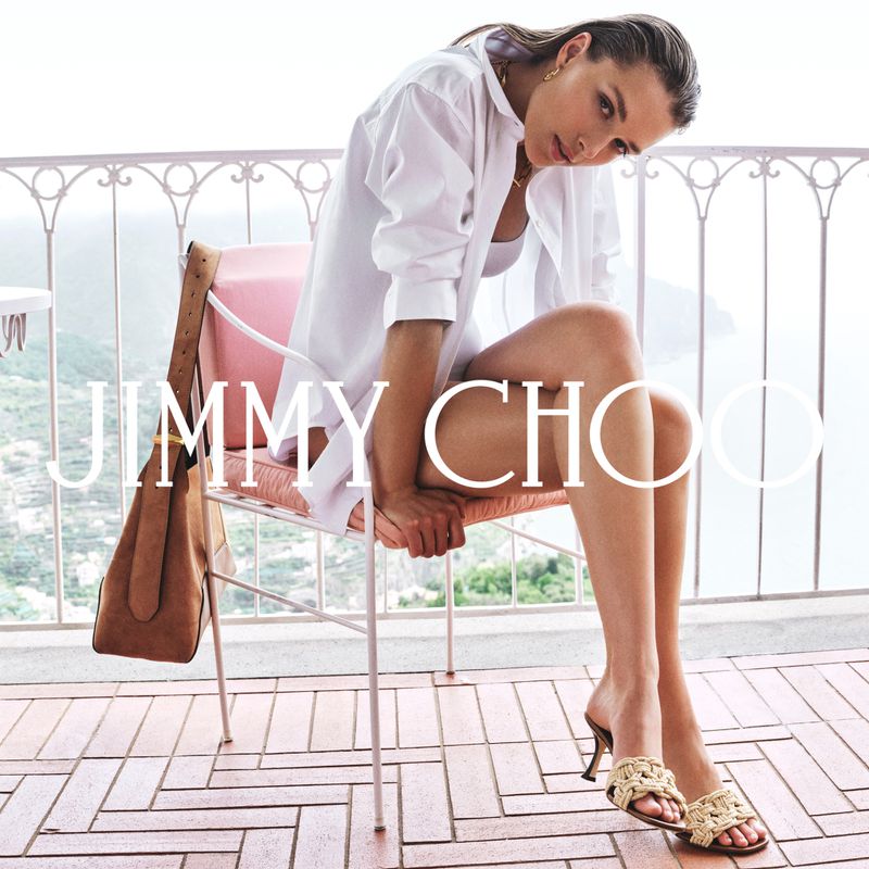 A model sits on a chair wearing a white shirt, jewelry, and mules with a bag hanging from the chair against a balcony and greenery.
