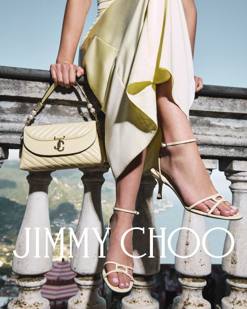 Model showcases Jimmy Choo cream leather stiletto sandals and a matching handbag with gold accents, posing outdoors with a scenic backdrop.