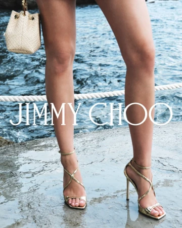 Close-up of a woman's legs and gold Jimmy Choo high heels standing on a stone surface, with a woven handbag and ocean background.