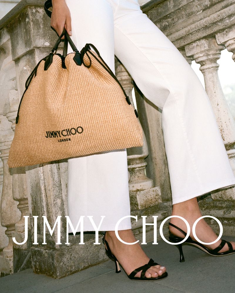 Jimmy Choo raffia tote bag and strappy heeled sandals are the focus against an outdoor stone terrace backdrop.