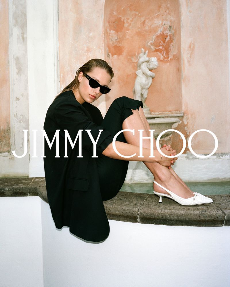 Fashion editorial shot of a woman in a black blazer and pants sitting in front of a statue wearing sunglasses and white heels.
