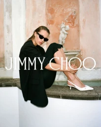 Model in black blazer and sunglasses seated near a fountain with white slingback heels for Jimmy Choo Bon Bon campaign
