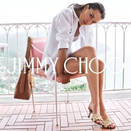 Model seated on a pink chair wearing a white shirt and woven sandals with a brown handbag for Jimmy Choo