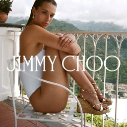 Model wearing a white swimsuit and metallic wedge sandals seated on a white balcony overlooking lush green mountains.