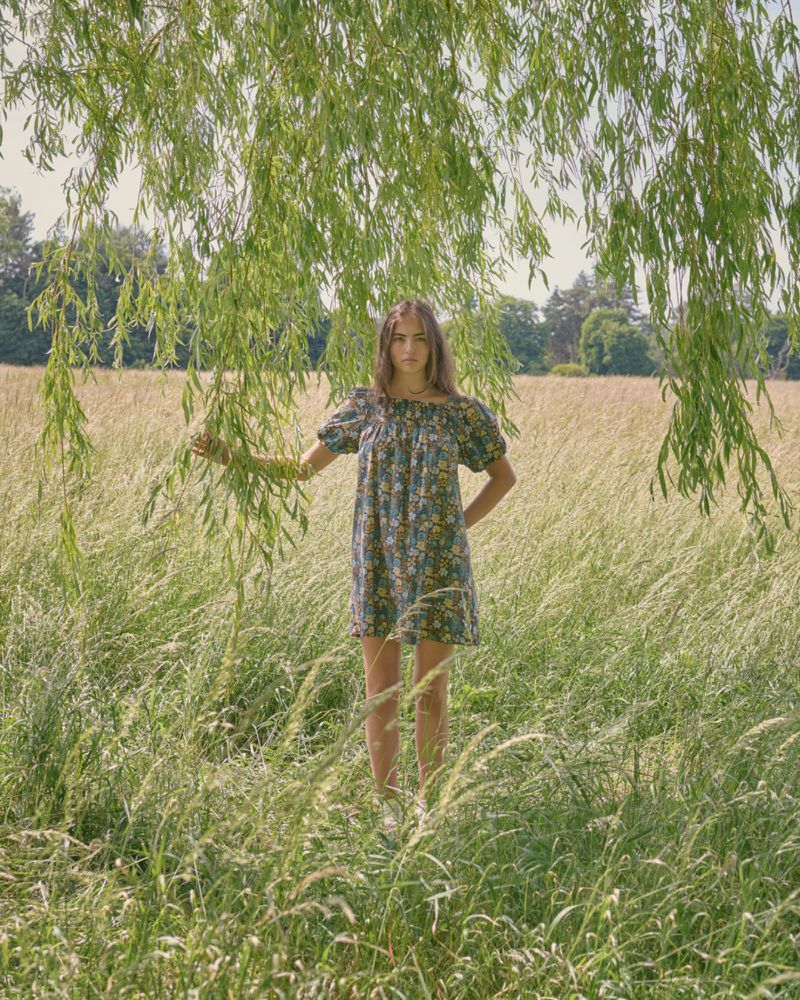 Model Stella Jones in a floral dress stands under hanging willow branches in a sunlit meadow of tall grass.