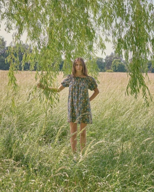 Model Stella Jones in a floral dress stands under hanging willow branches in a sunlit meadow of tall grass.