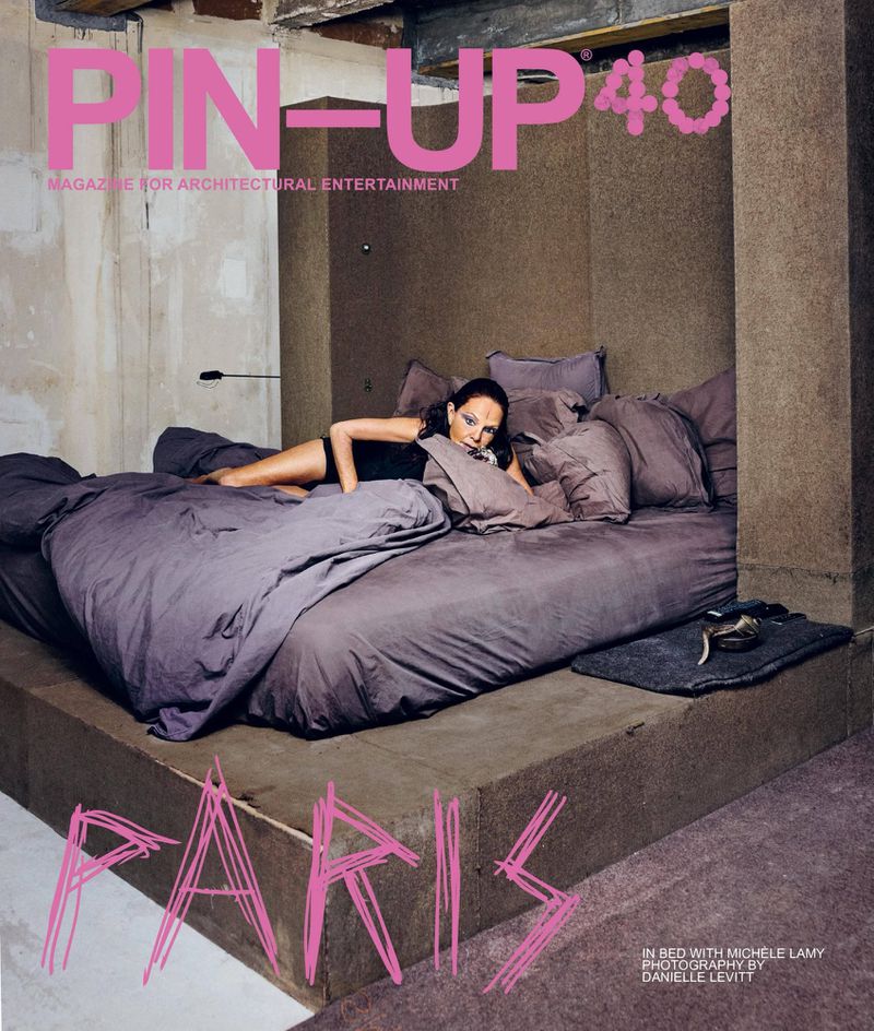 Michèle Lamy reclining in a large platform bed with grey bedding inside a textured architectural alcove for PIN-UP Magazine.