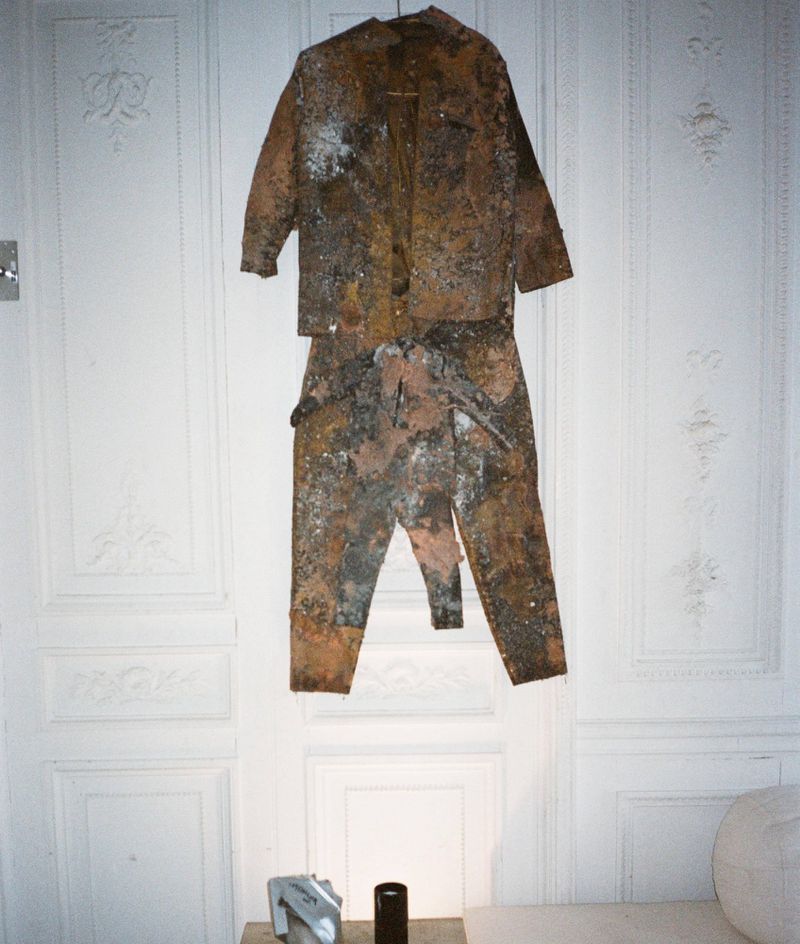 Weathered and oxidized brown suit hanging flat against a white wall with ornate classical decorative moulding.