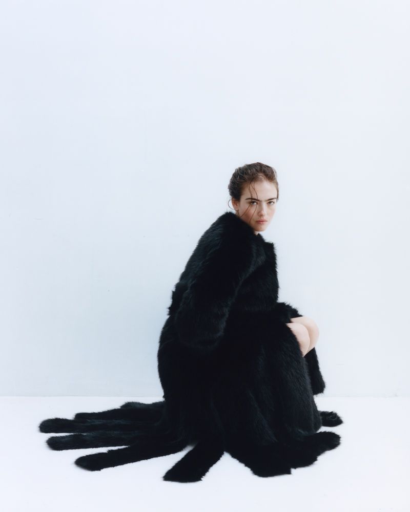 Model Stella Jones crouching in a voluminous black fur coat with trailing fringe against a seamless white studio background.