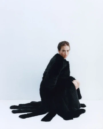 Model Stella Jones crouching in a voluminous black fur coat with trailing fringe against a seamless white studio background.