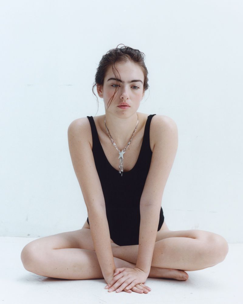 Model Stella Jones seated cross-legged wearing a black sleeveless bodysuit and star pendant necklace against a white background.
