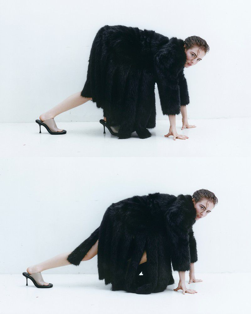 Model Stella Jones crouching in an oversized black textured coat and black high-heeled mules against a bright white seamless background.
