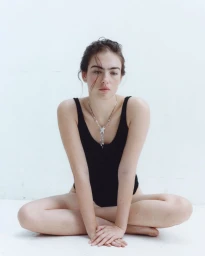 Model Stella Jones seated cross-legged wearing a black sleeveless bodysuit and star pendant necklace against a white background.