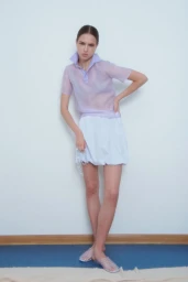 Model in a sheer lavender polo shirt and white bubble skirt standing against a neutral wall with hand on hip.