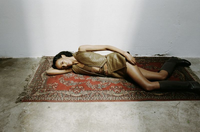 Model Amina Ahmed lying on a patterned red rug wearing an olive quilted leather two-piece outfit and knee-high boots.