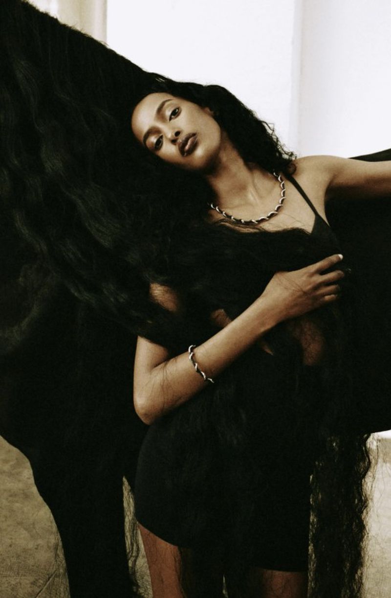Model Amina Ahmed posing in a black dress with sweeping long dark hair and metallic chain jewelry.