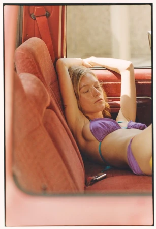 Woman with blonde hair wearing a purple bikini resting her arms behind her head on the red upholstered seat of a vintage car.