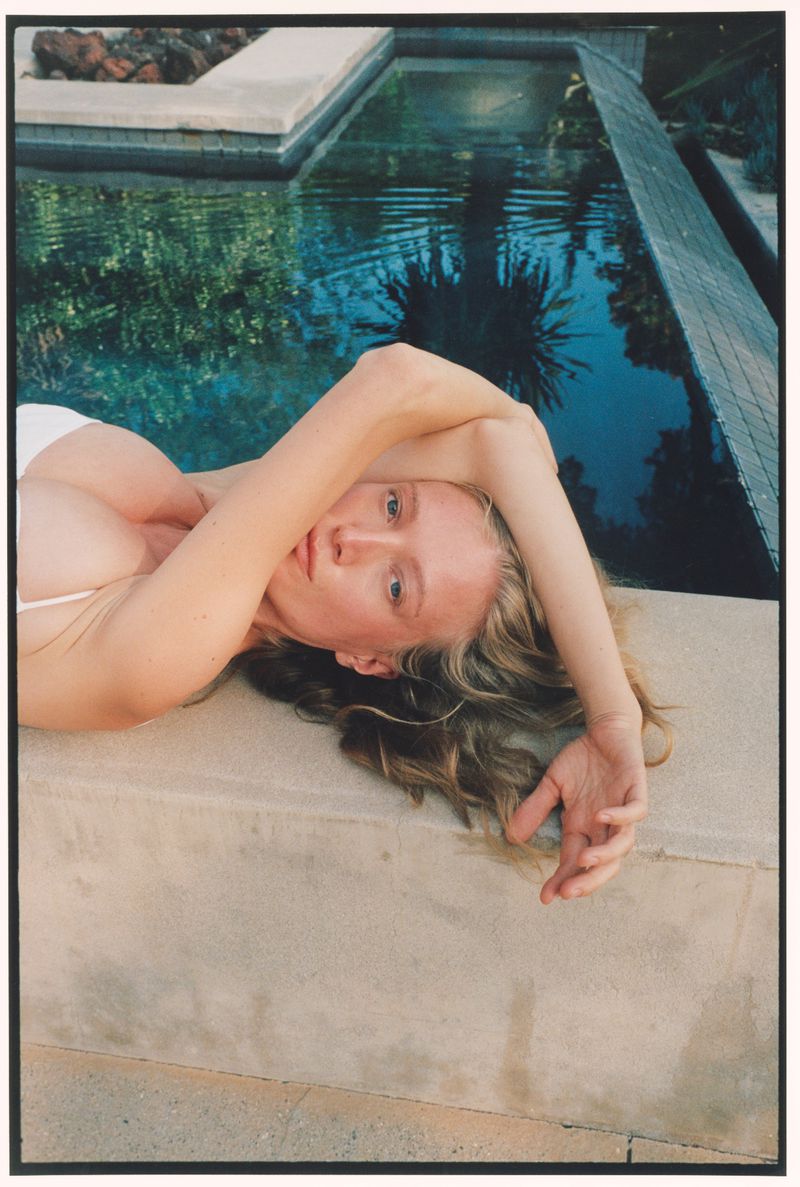 Woman in a white bikini top reclining along the concrete edge of an outdoor swimming pool with clear water reflecting palm trees.